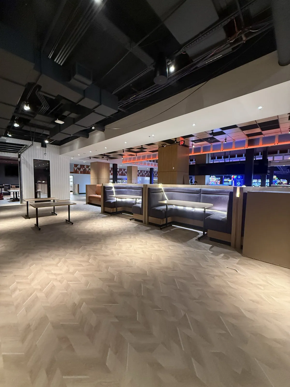 Row of grey upholstered booths with light-wood bases and end panels, wood-clad columns and arcade games behind