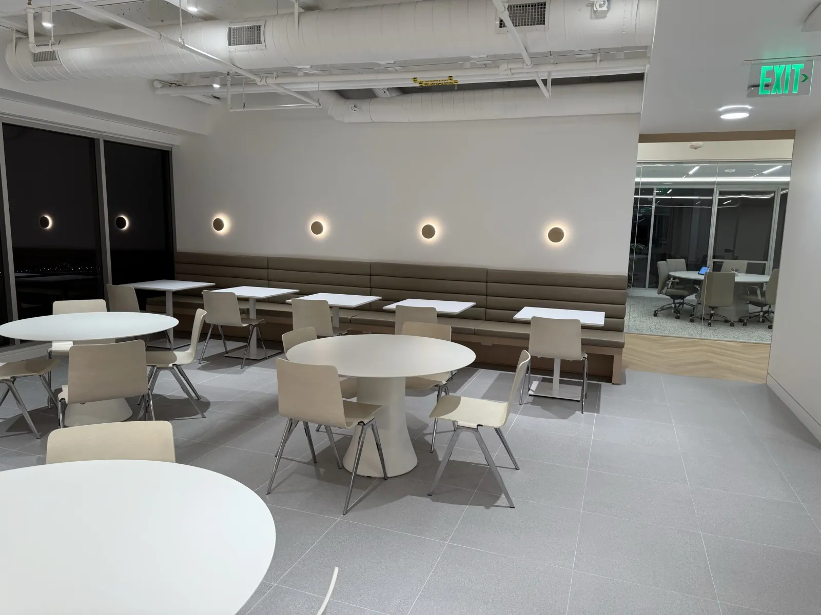 Long upholstered banquette along a break-area wall under round wall lights, with tables and chairs
