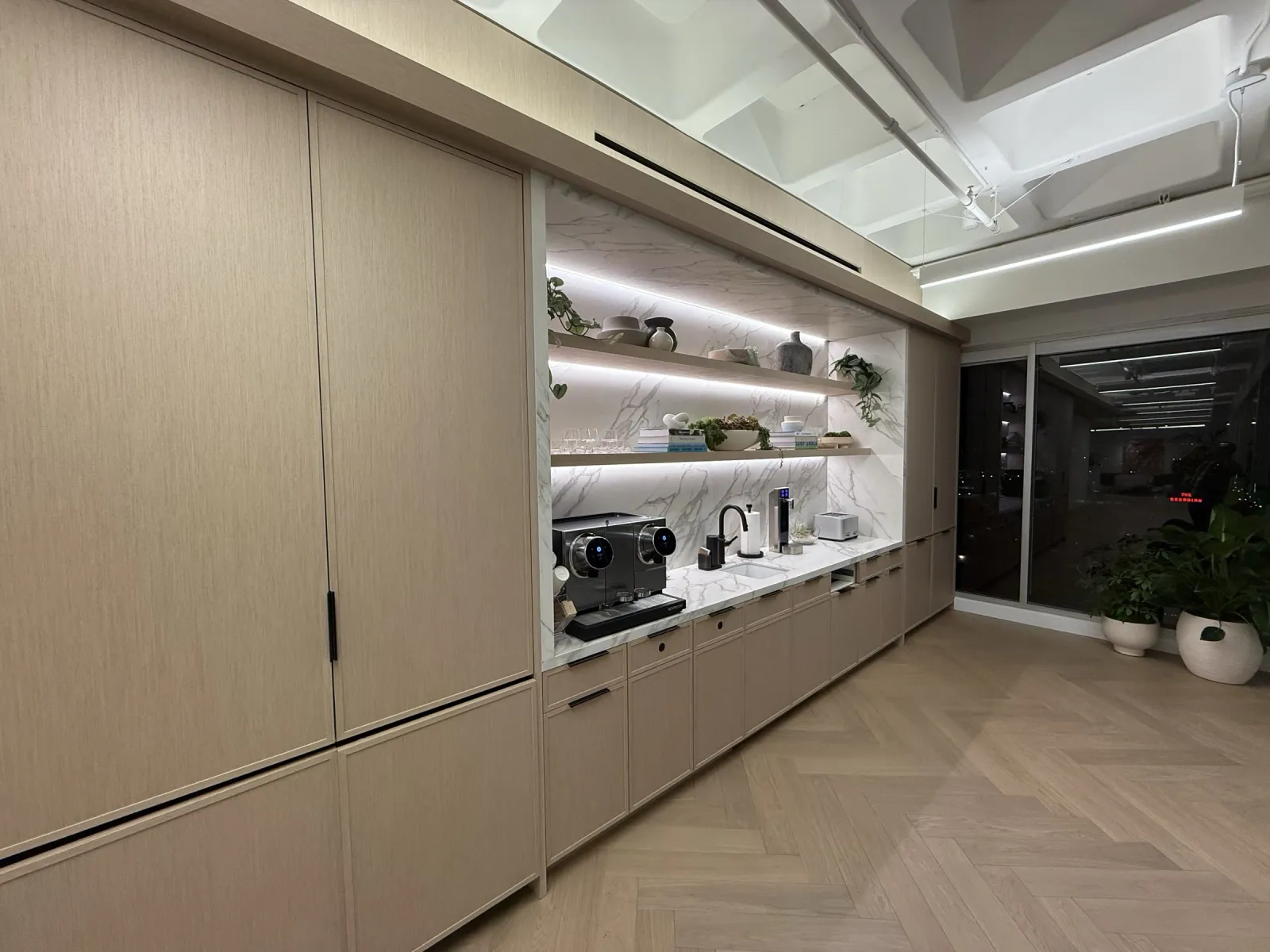Long wood-grain storage wall with a lit open-shelf niche over a marble-look coffee counter