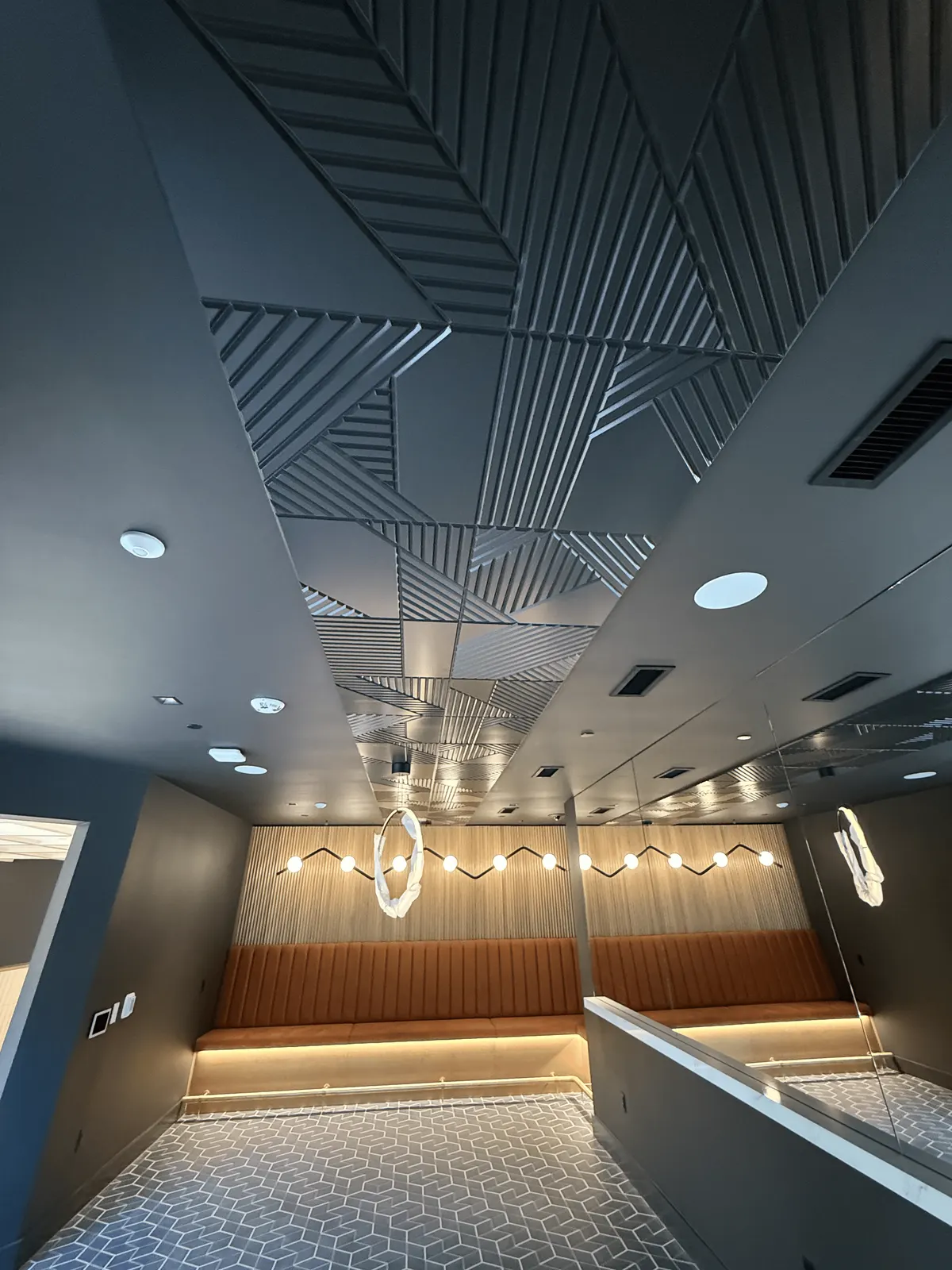 Game lounge ceiling of dark textured panels with angled slat patterns above a banquette and fluted wall