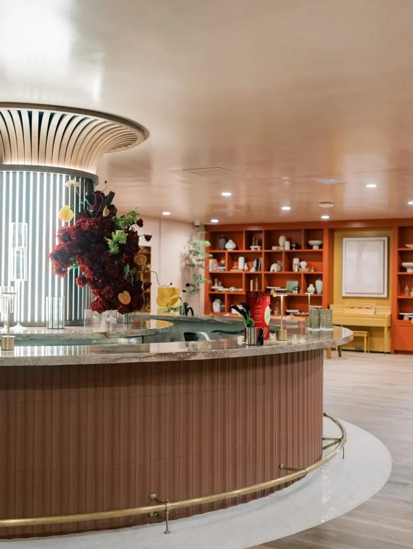 Circular bar at The Linden Club with a stone top and fluted base; the painted library built-in and piano are behind