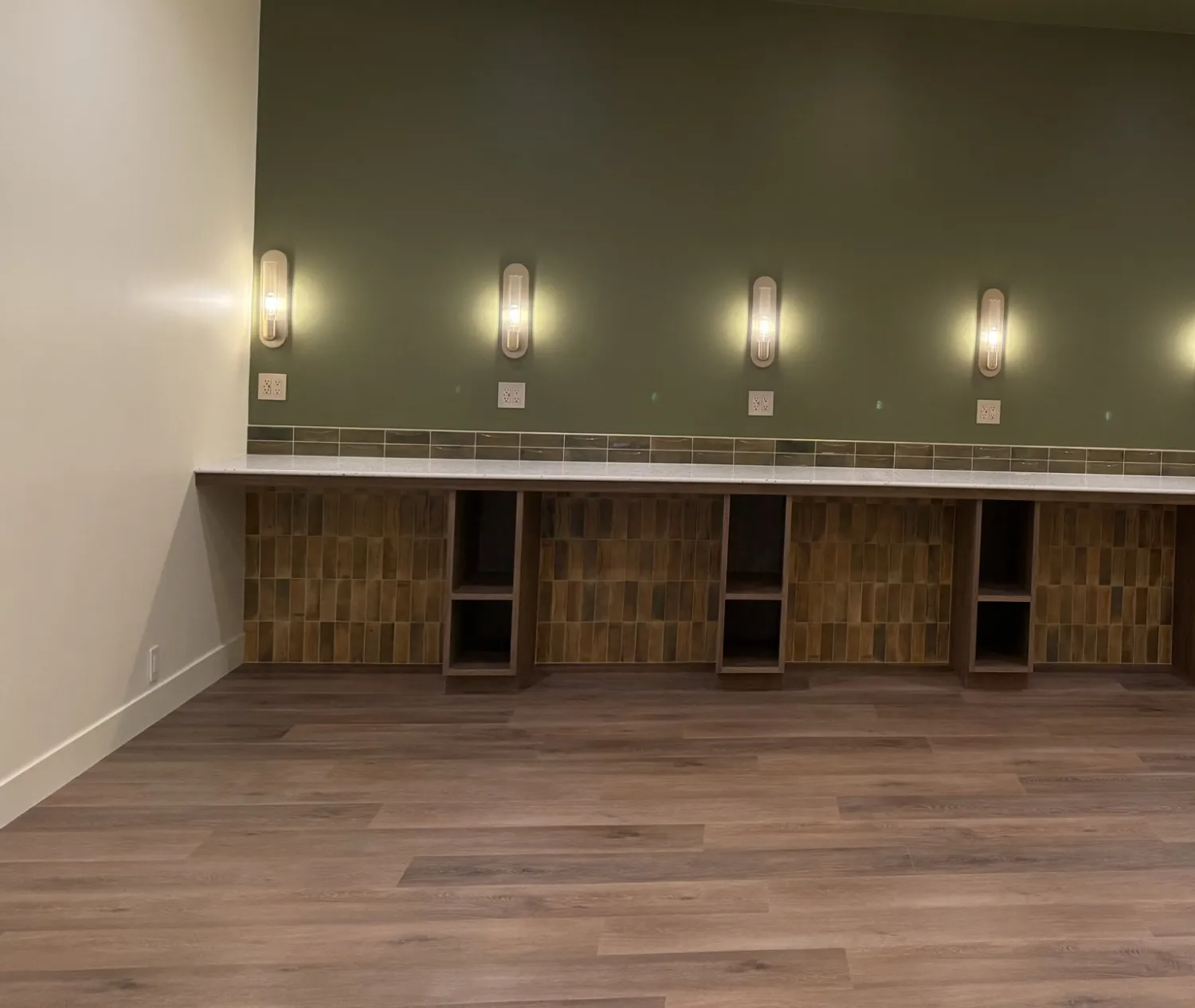 Long vanity counter on walnut base cabinets with open cubbies, under a row of wall sconces on a green wall