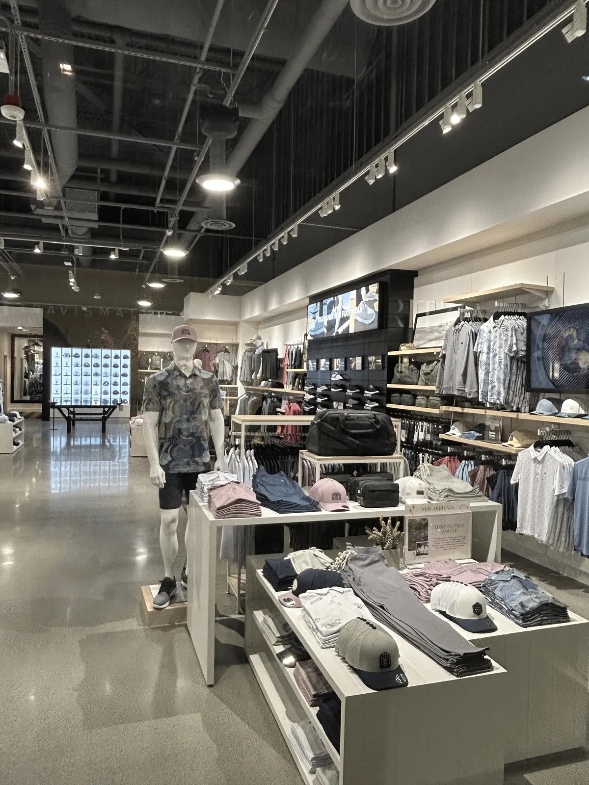 Light wood wall shelves holding caps and bags beside white merchandise tables in the TravisMathew store