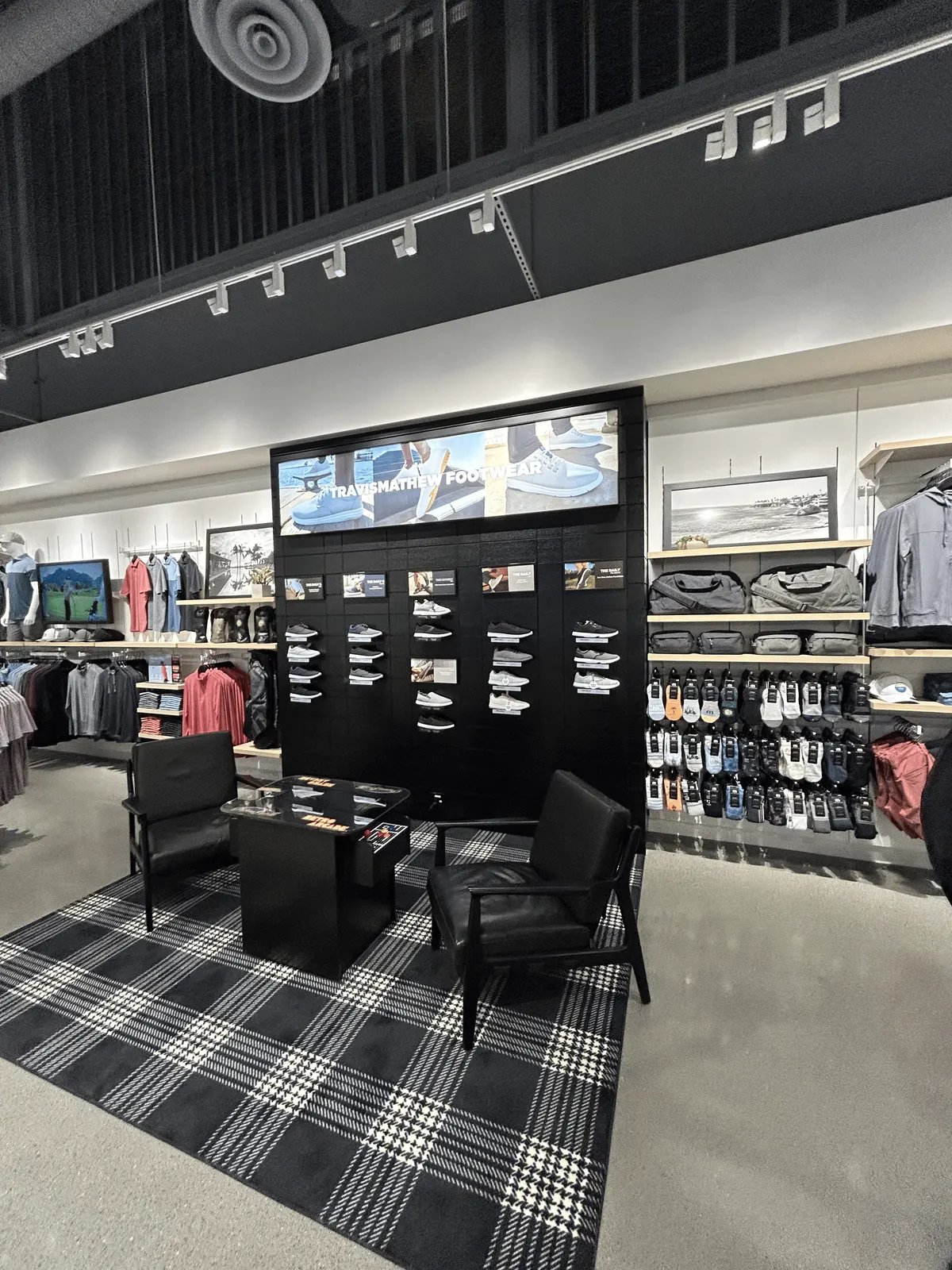 Black paneled footwear display wall with shoe shelves, flanked by light wood shelving for bags and socks